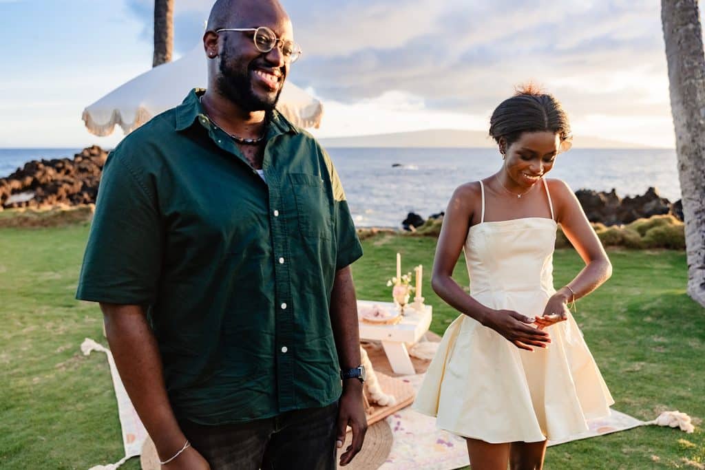 Ernest smiling and laughing as Gloria is smiling and looking down at her new engagement ring. The picnic setup glowing in the sunset behind them.