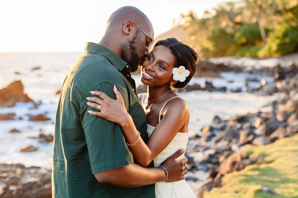 Ernest and Gloria embracing on the coast of Maui as Ernest gives Gloria a kiss on the cheek and she smiles and holds onto his shoulders.