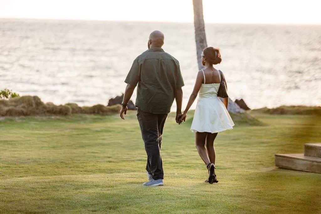 A couple holding hands and walking towards the beach. The man is wearing a button up tee and slacks, the woman is wearing a short white dress and black heels.