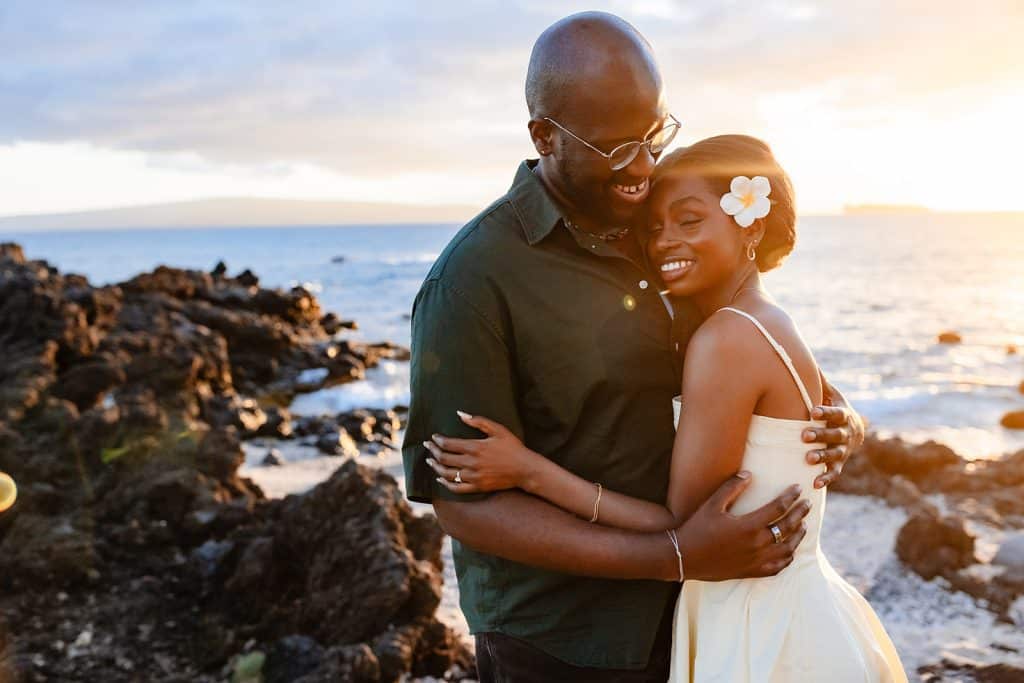Ernest and Gloria hugging on the rocky Maui coast as the sun is setting behind them. Ernest is smiling and looking down at Gloria as she leans into him with her eyes closed and a smile on her face.
