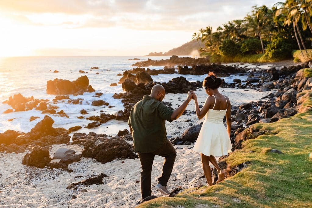 Ernest stepping onto the sand and holding Gloria's hand as he guides her down to the beach as the sun is setting on the water.