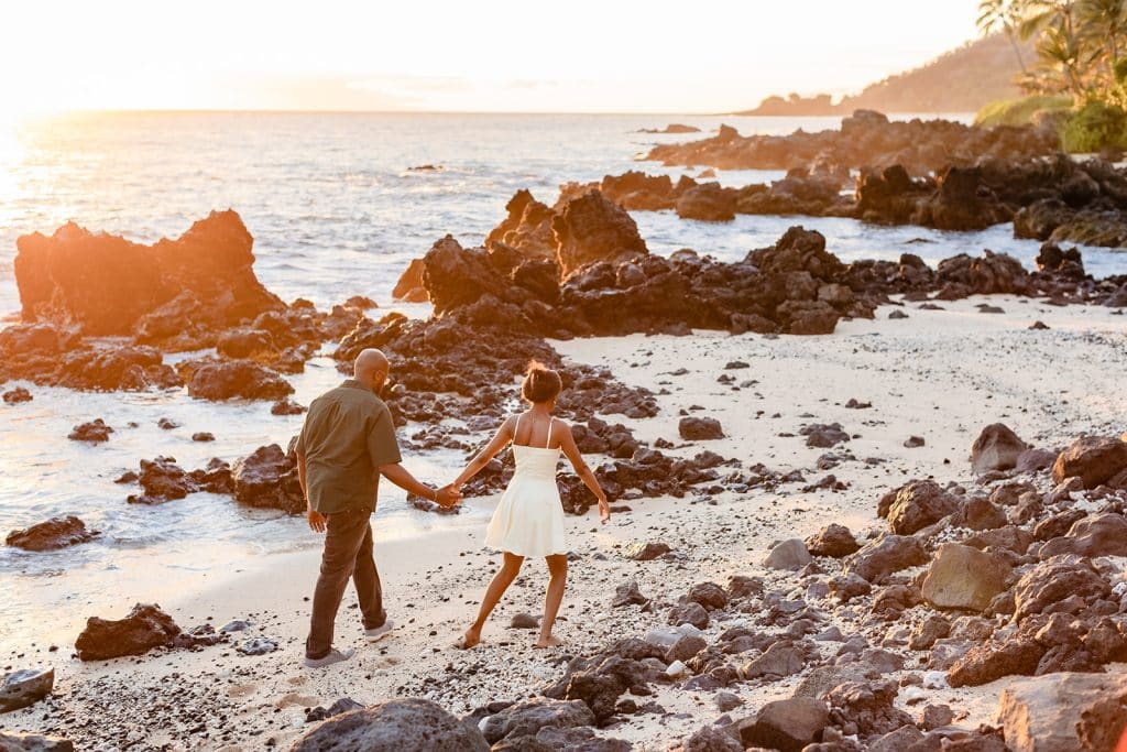 Ernest and Gloria walking in the sand and holding hands down the coast of the rocky beach as the sun sets.