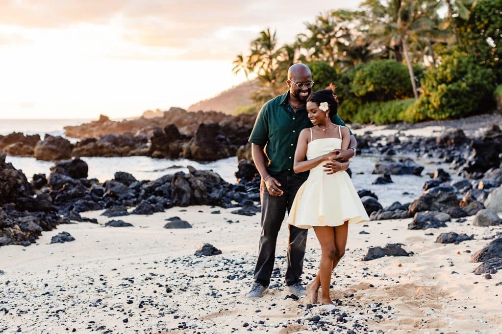 Ernest standing behind Gloria on the beach and holding her around the waist as she leans into him and grabs his hand. Ernest is smiling at Gloria as she smiles into him